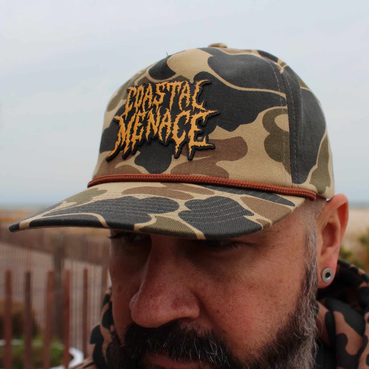 Person wearing a camouflage hat with "Coastal Menace" text, standing outdoors by the beach.