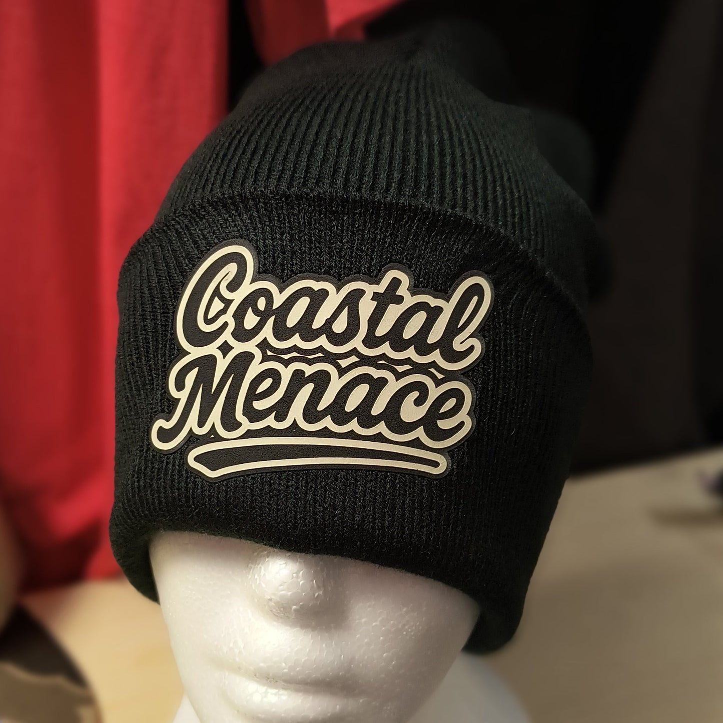 close up of retro script Coastal Menace patch on a black beanie