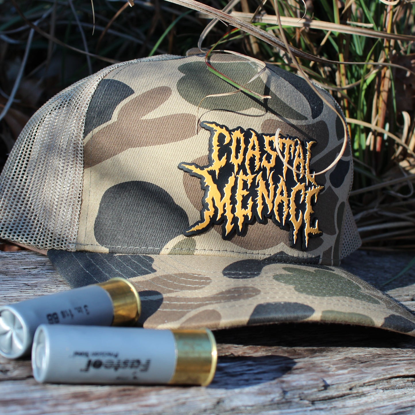 Camouflage cap with 'Coastal Menace' logo on a wooden surface with shotgun shells.