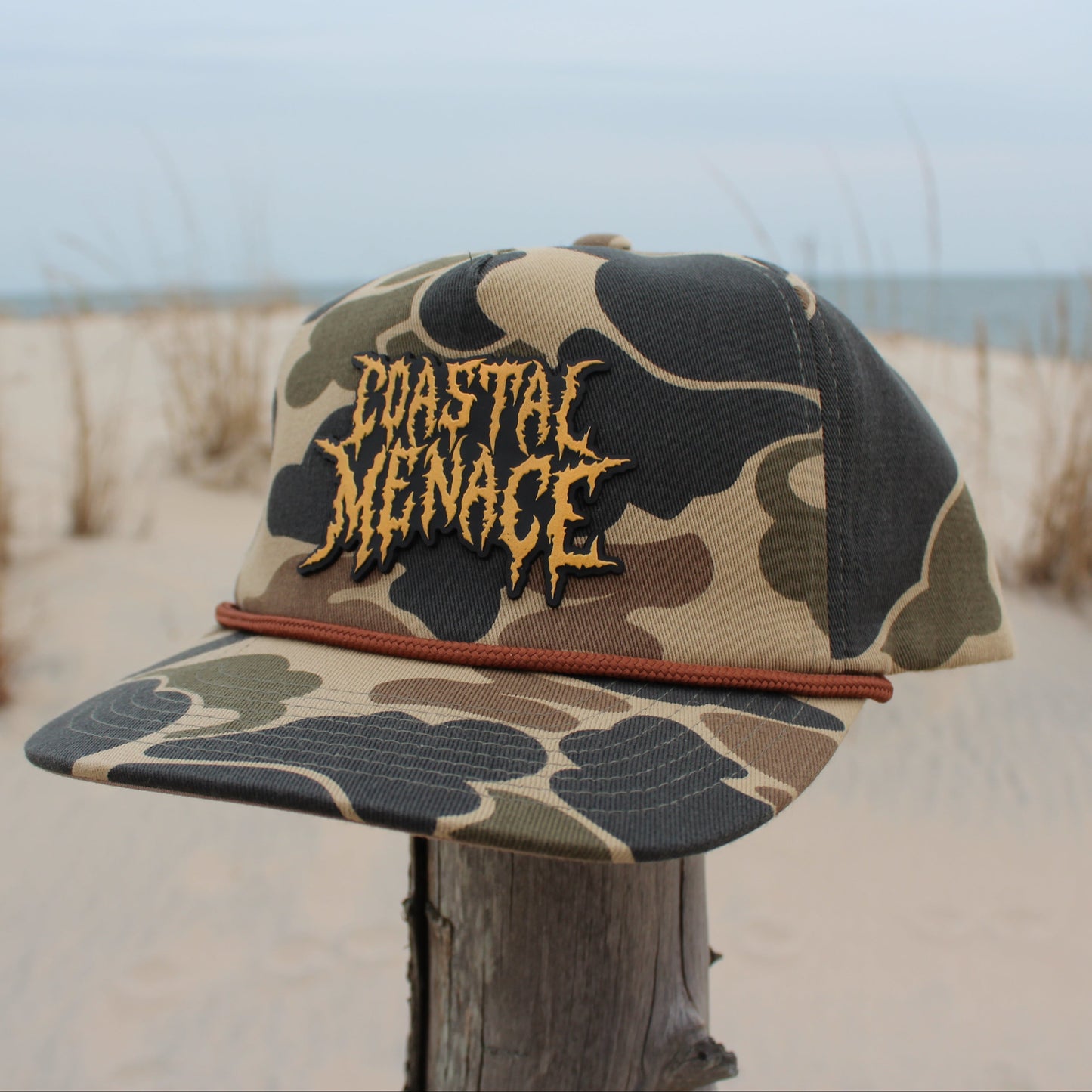 Camouflage cap with 'Coastal Menace' text on a wooden post with beach background