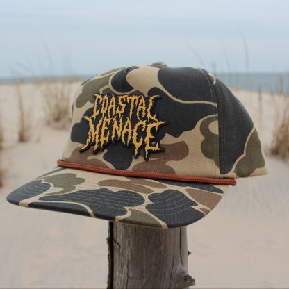 Camouflage cap with 'Coastal Menace' text on a wooden post with beach background