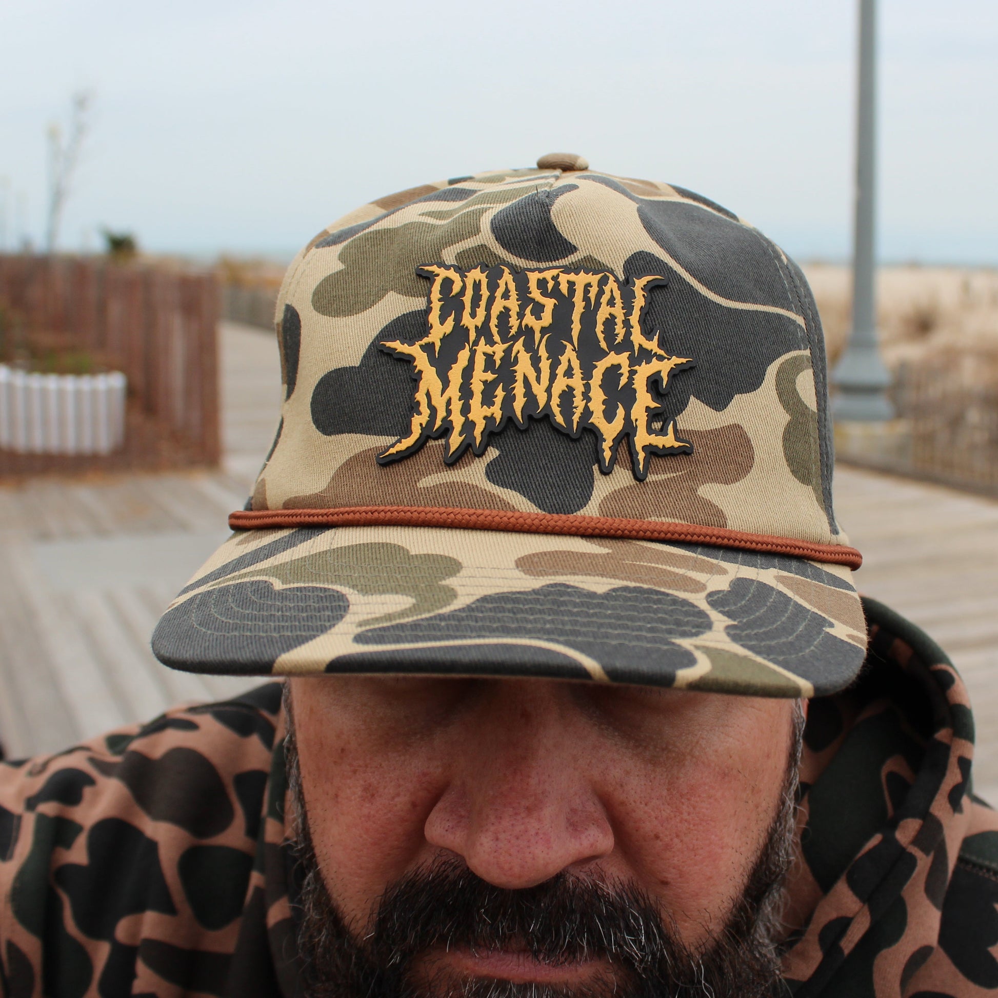 Person wearing a duck camouflage cap with 'Coastal Menace' text, standing on a wooden boardwalk.