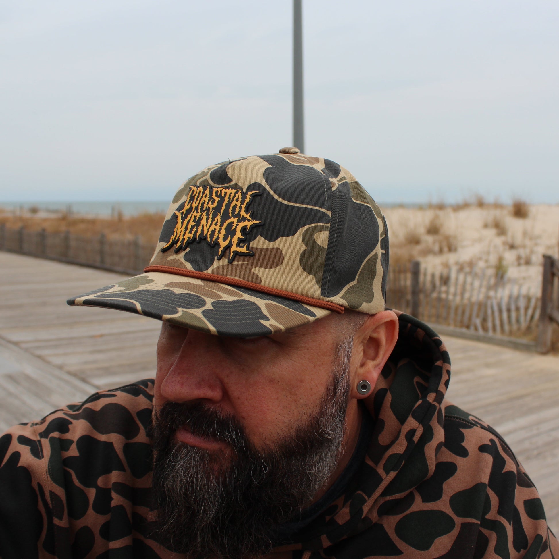 Person wearing a duck camouflage cap with 'Coastal Menace' text on a boardwalk.