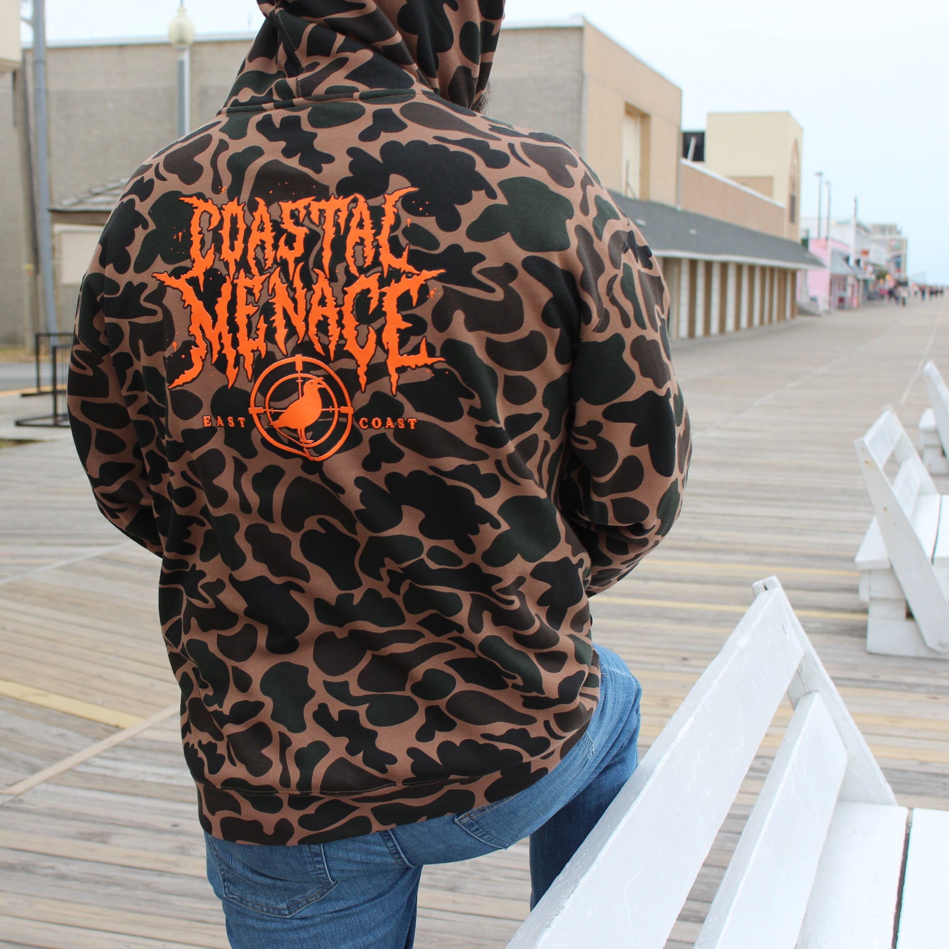 Person wearing a duck camo print hoodie with 'Coastal Menace' branding on a boardwalk.