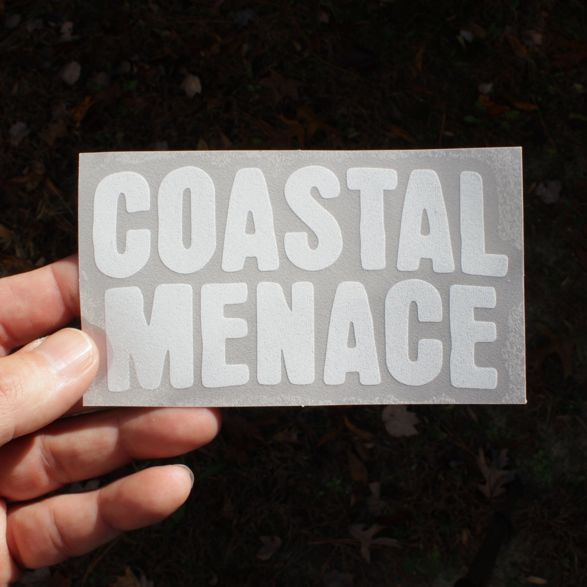 Hand holding a clear decal with 'COASTAL MENACE' text against a dark background