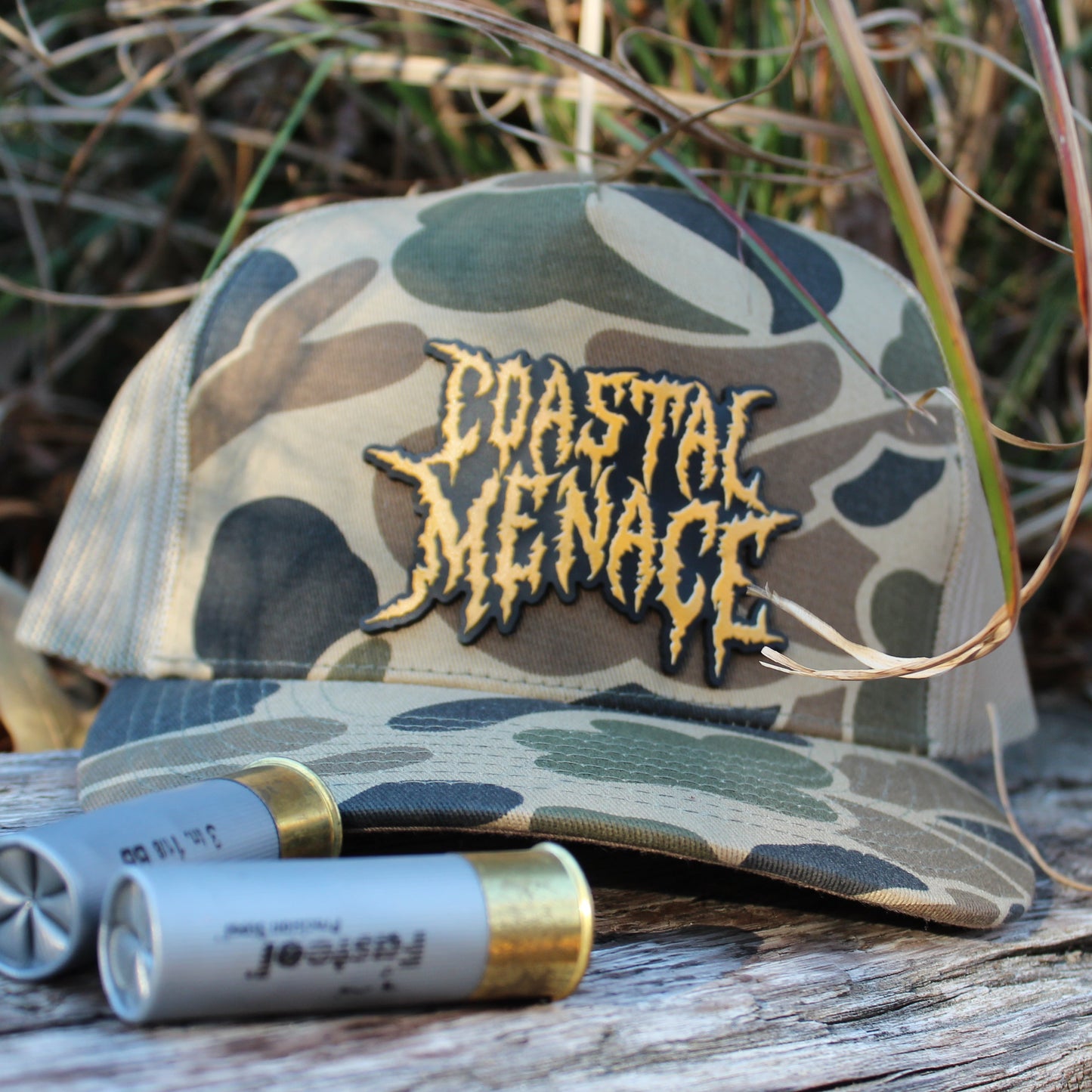 Camouflage cap with 'Coastal Menace' logo on a log with shotgun shells, surrounded by marsh grass.