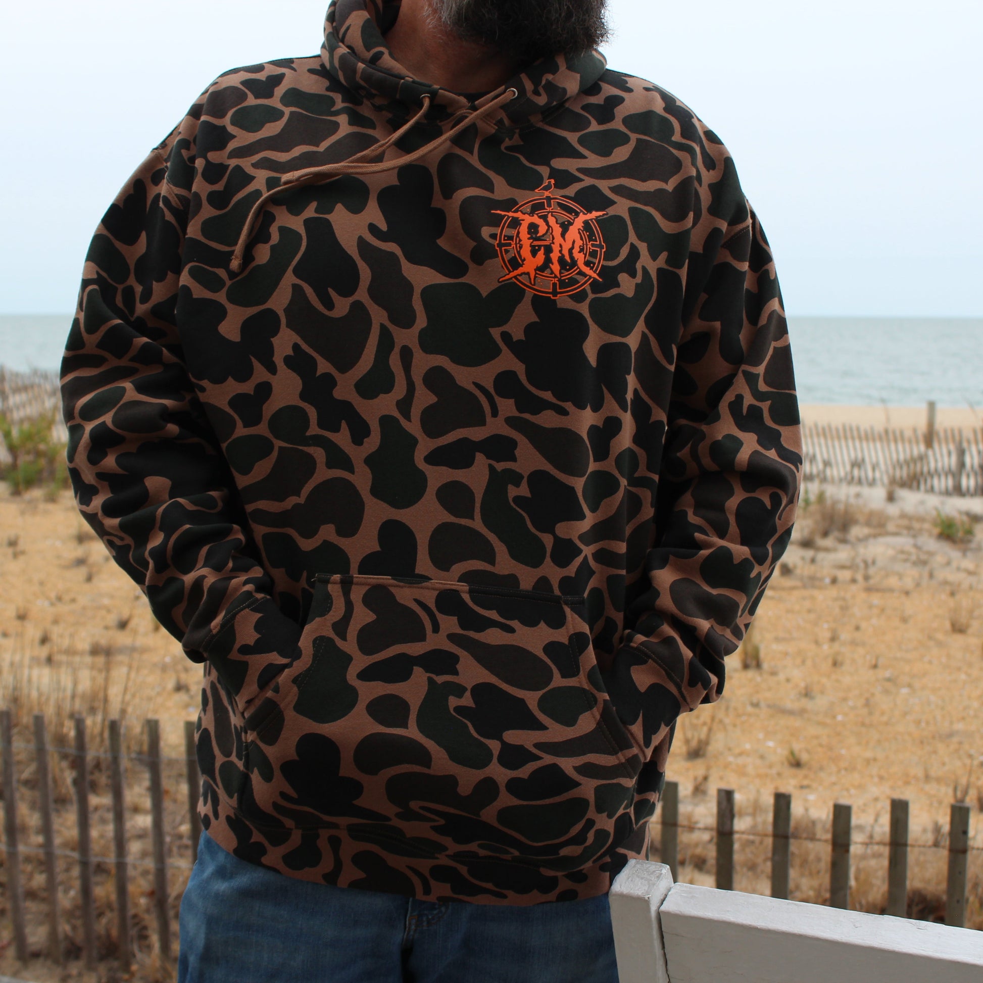 Person wearing a camo print hoodie with a logo, standing on boardwalk.