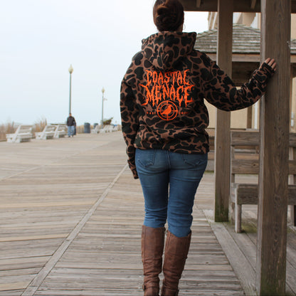 Person wearing a camo print hoodie with 'Coastal Menace' text on a wooden boardwalk by the beach