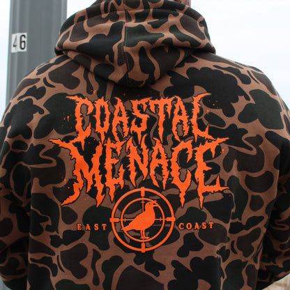 Camouflage hoodie with 'Coastal Menace East Coast' text and logo on the back, worn by a person by the beach