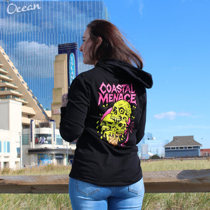 Person wearing a black hoodie with 'Coastal Menace' design, standing by a wooden railing with buildings and blue sky in the background.