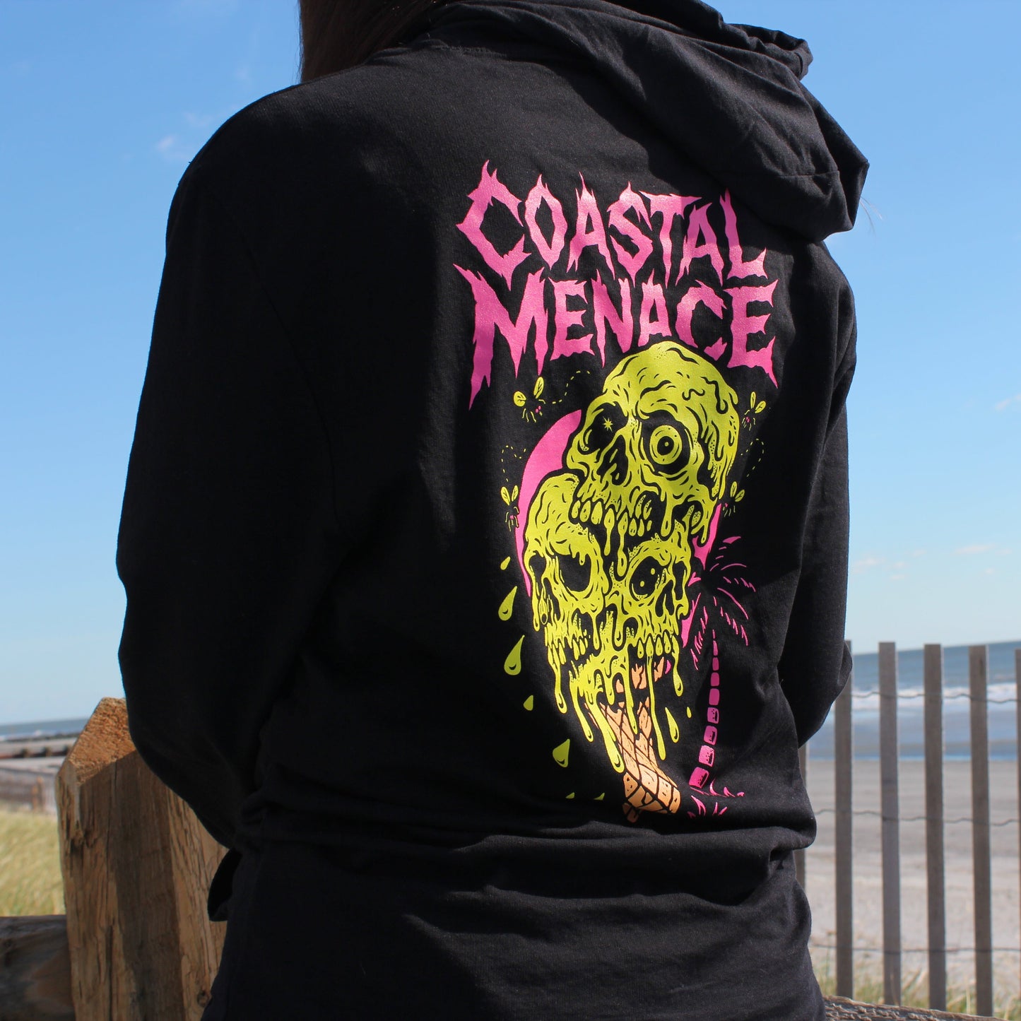 Black hoodie with 'Coastal Menace' design on the back, worn by a person at a beach.