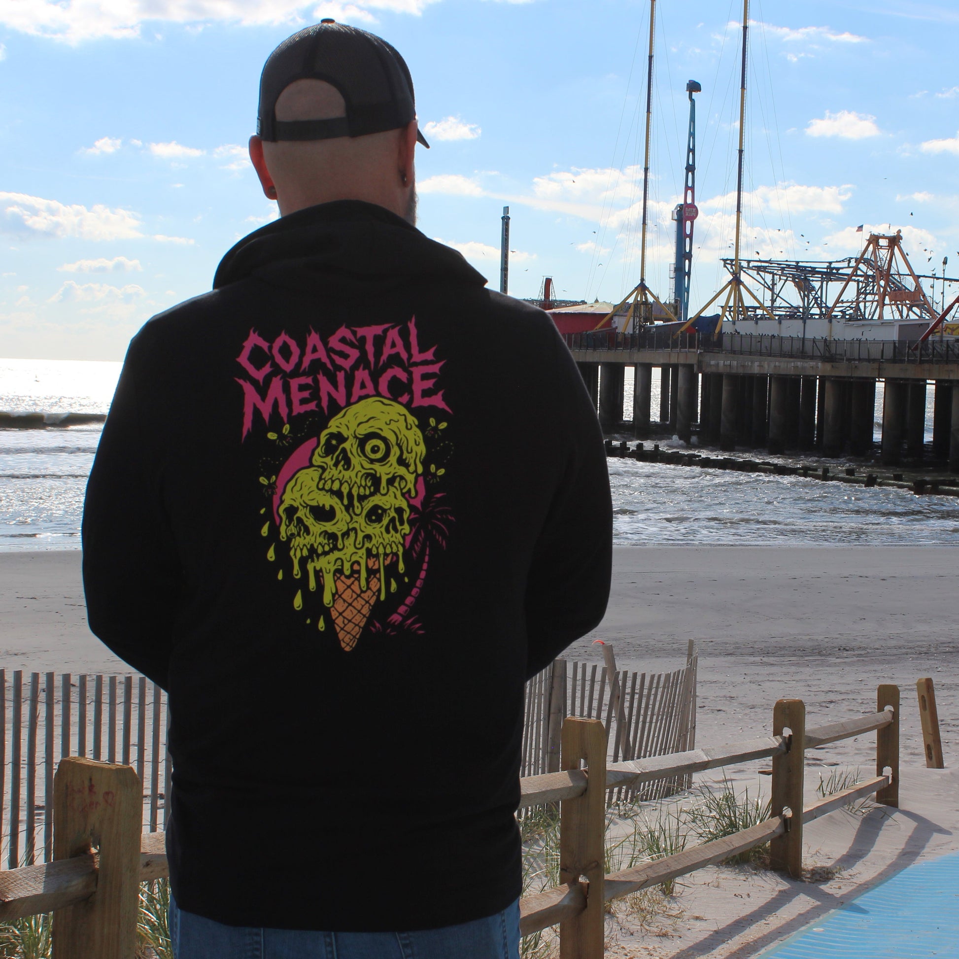Person wearing a hoodie with 'Coastal Menace' design at a beach with a pier and Ferris wheel in the background.