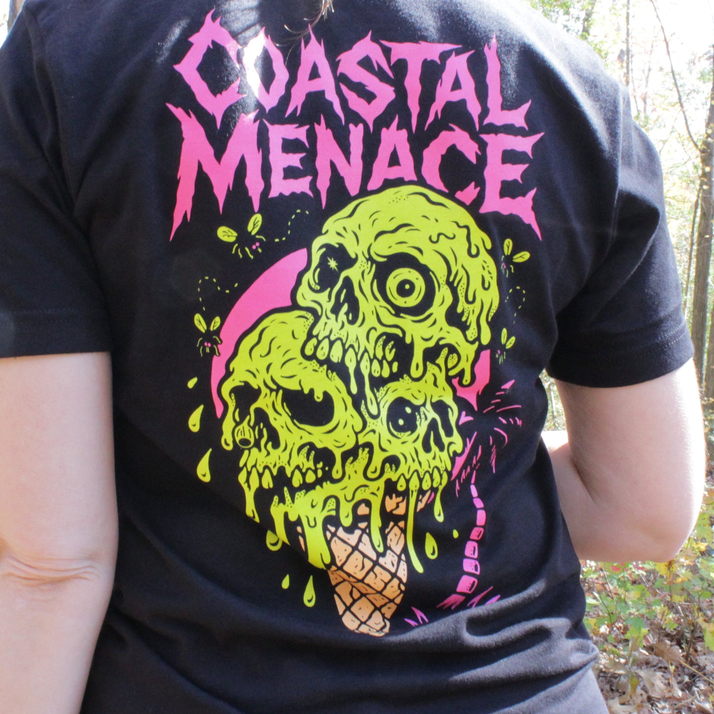 Person wearing a black t-shirt with a colorful skull design and 'Coastal Menace' text in a forest setting.
