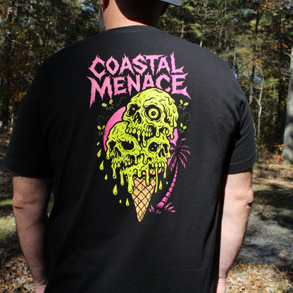 Person wearing a black t-shirt with 'Coastal Menace' colorful design in a forest setting