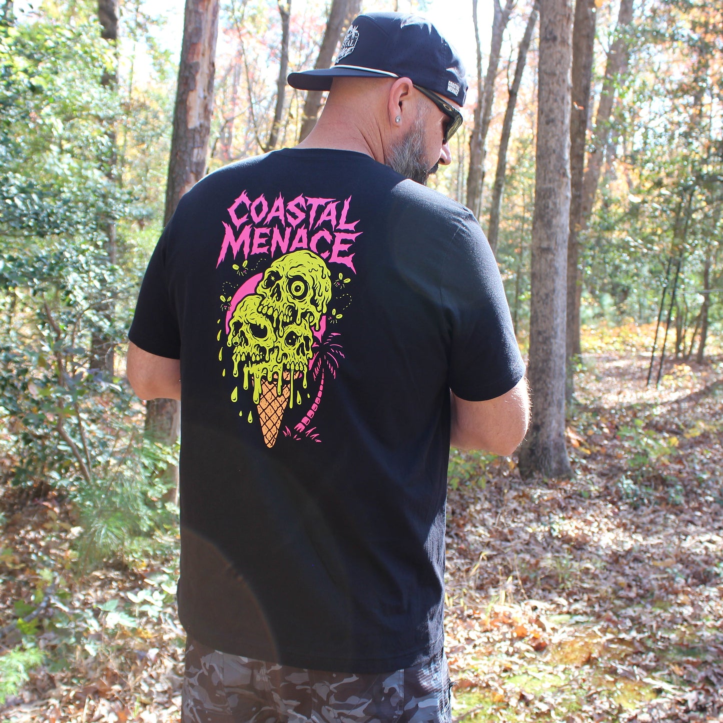 Man wearing a black t-shirt with colorful skull 'Coastal Menace' design in the woods