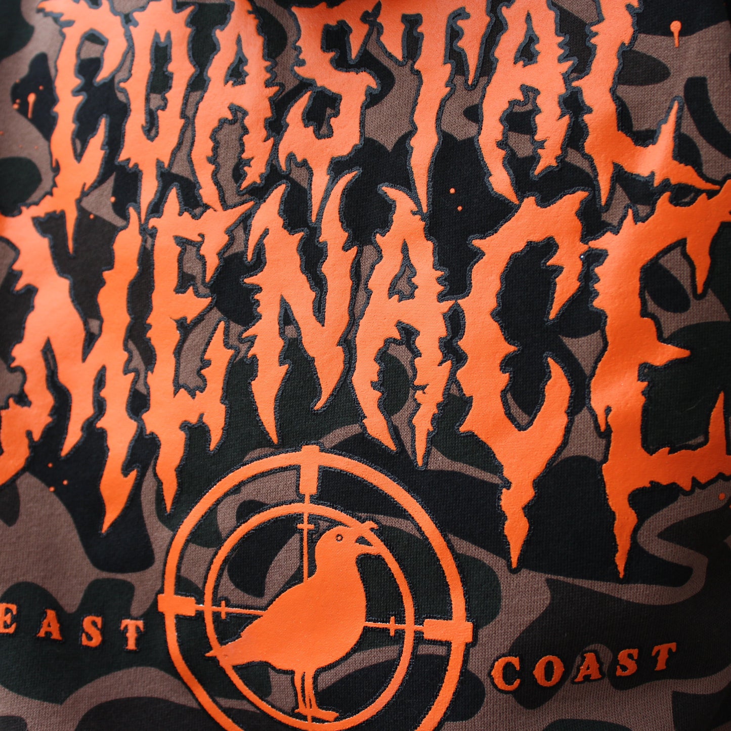 Close-up of a bright orange Coastal Menace printed logo on a camo hoodie.
