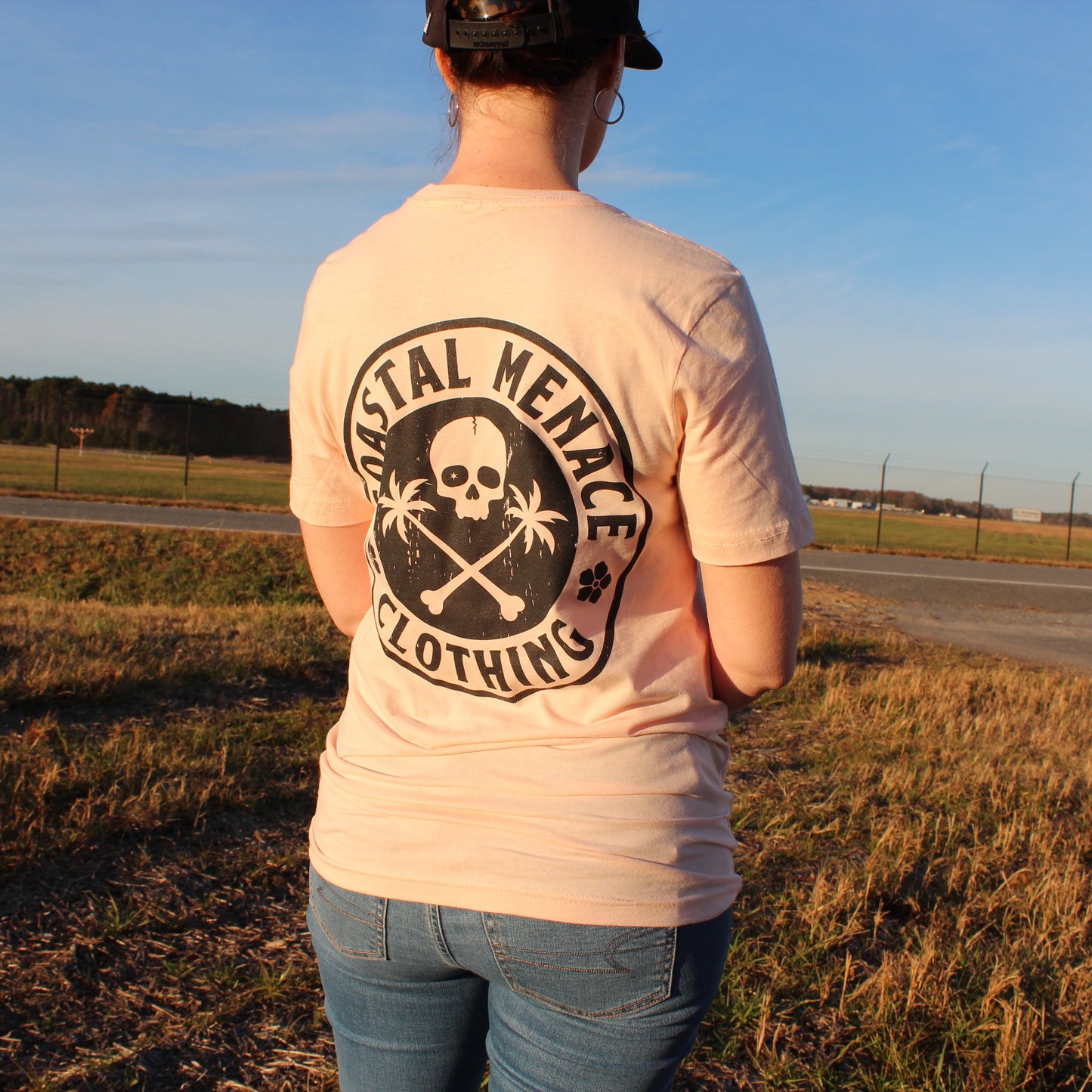 Back view of women’s Coastal Menace Base Threat pink t-shirt featuring large skull-and-palms logo.