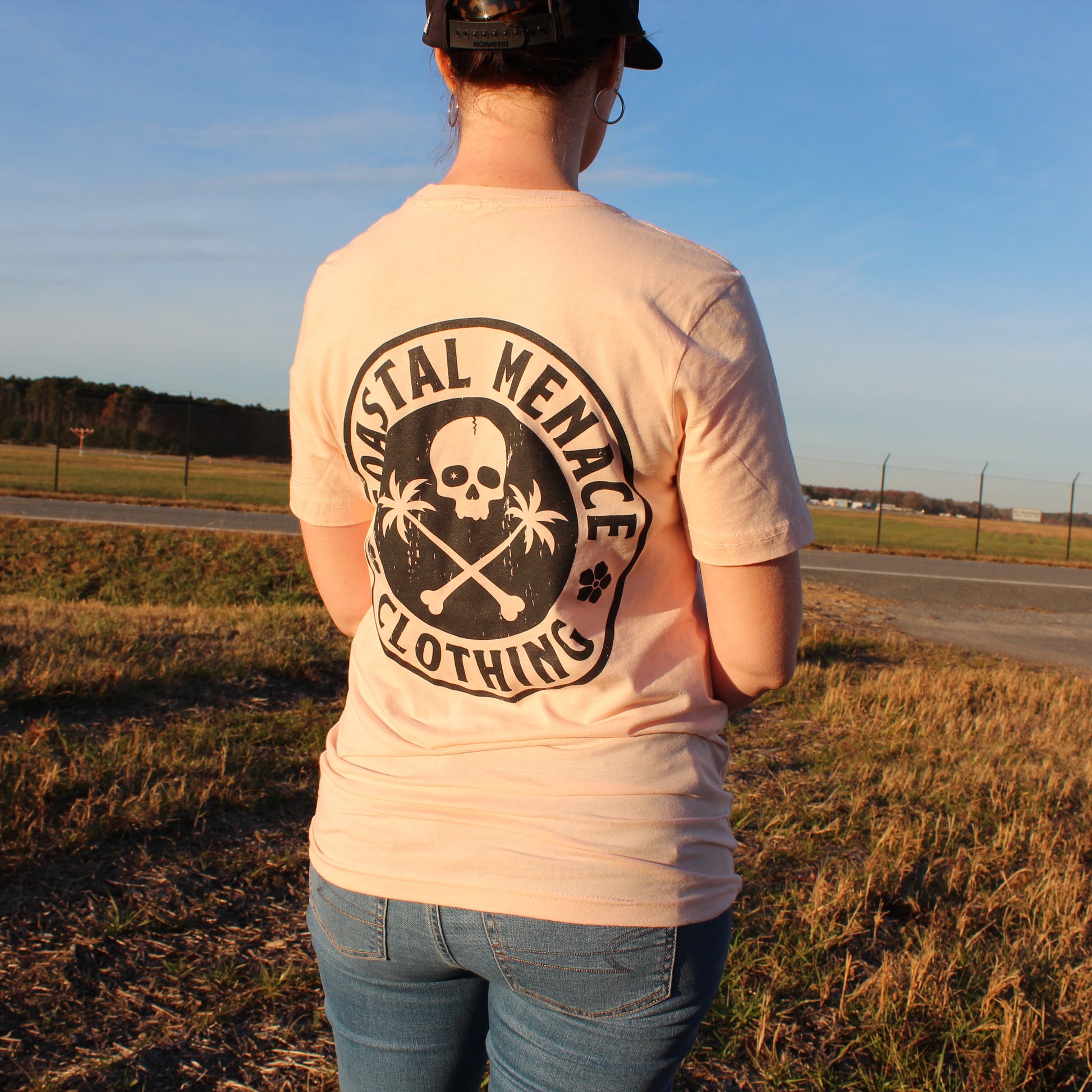 Back view of women’s Coastal Menace Base Threat pink t-shirt featuring large skull-and-palms logo.