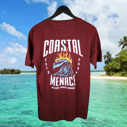 Back view of maroon Coastal Menace Hot Breaker tee featuring large flaming wave graphic and bold lettering, shown in front of a tropical shoreline scene