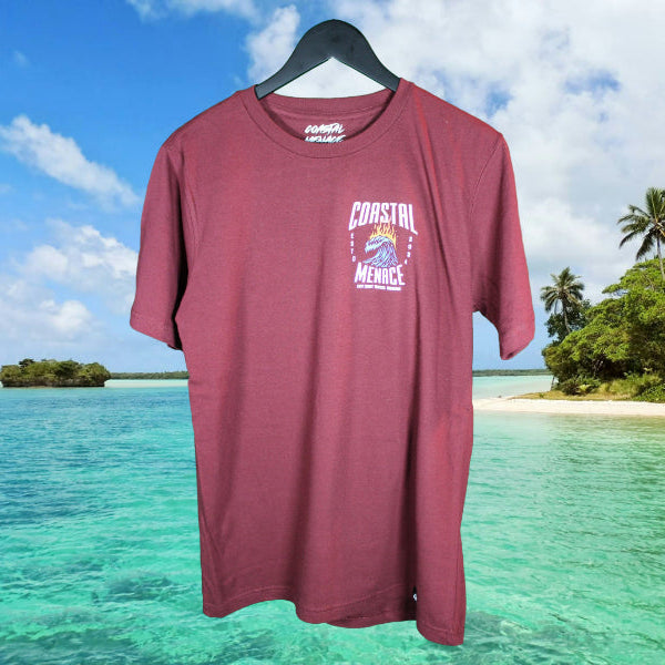 Maroon Hot Breaker T-shirt from Coastal Menace with small flaming wave logo on the left chest, photographed against a tropical beach backdrop