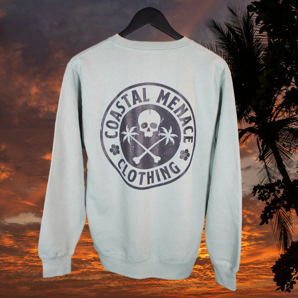 Seafoam Coastal Menace crewneck showing large back graphic with skull, palm trees, and crossed bones, photographed on a tropical sunset backdrop