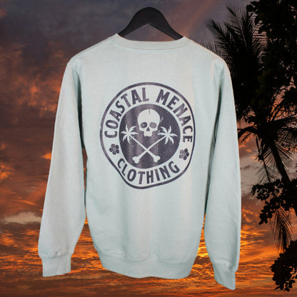 Seafoam Coastal Menace crewneck showing large back graphic with skull, palm trees, and crossed bones, photographed on a tropical sunset backdrop