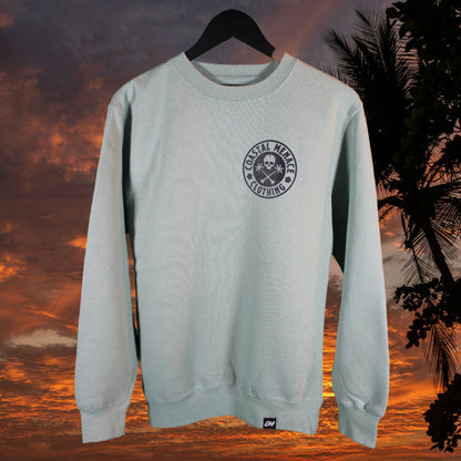 Seafoam crewneck sweatshirt with small Coastal Menace badge logo printed on the left chest, displayed against a tropical sunset background