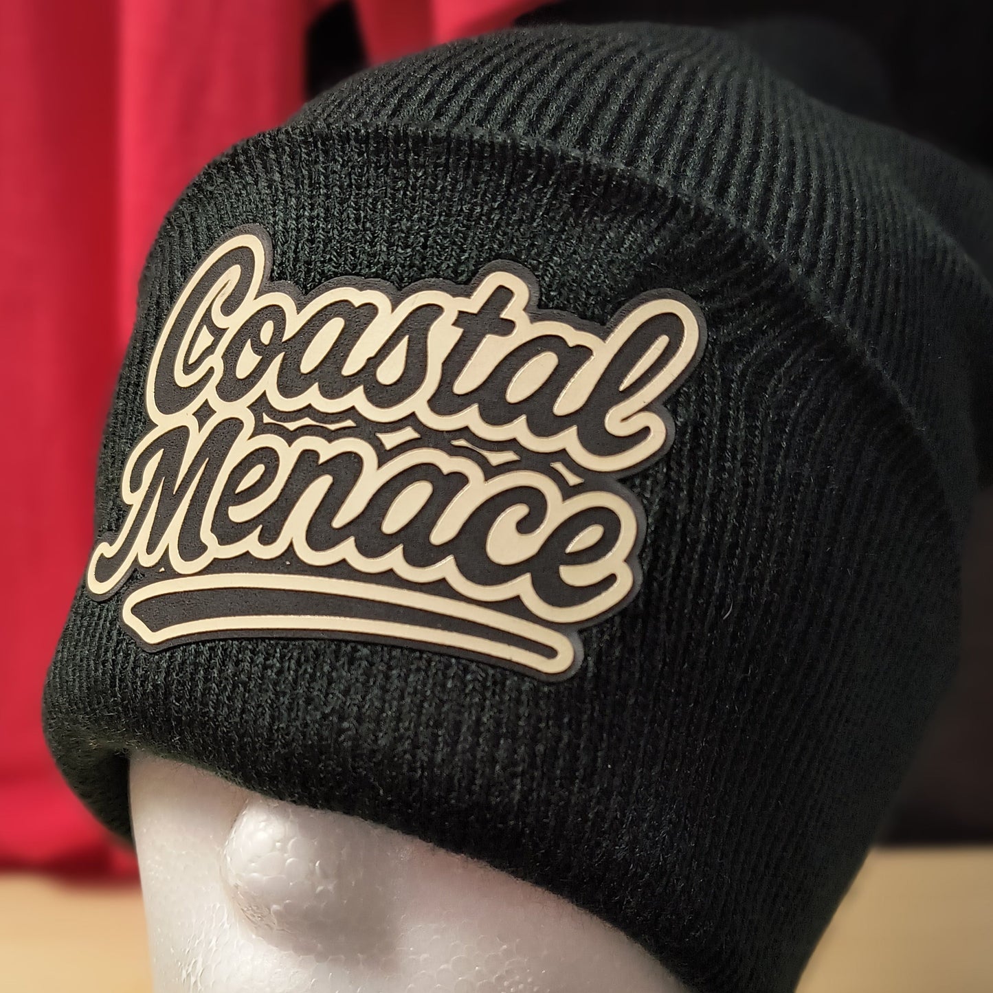 close up of retro script Coastal Menace patch on a black beanie