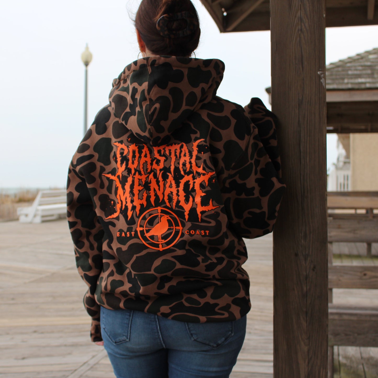 Person wearing a duck camo print hoodie with 'Coastal Menace' text on a wooden boardwalk by the beach.