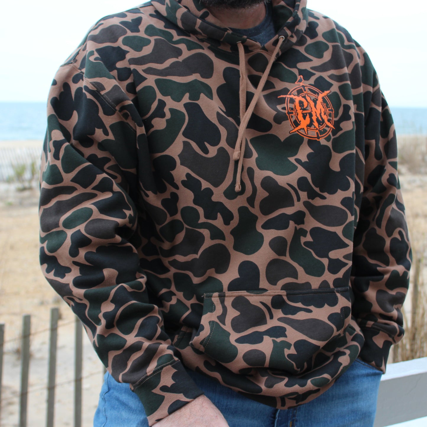 Person wearing a camouflage hoodie with a blaze orange logo, standing outdoors with a beach background.